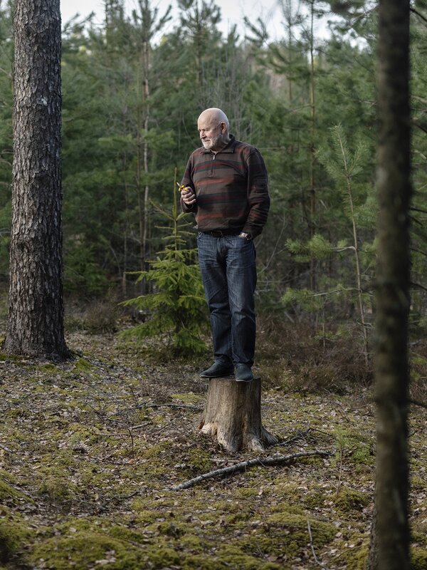 Composer Peteris Vasks stands on a tree stump in the woods in Latvia. Photo by Reinis Hofmanis.