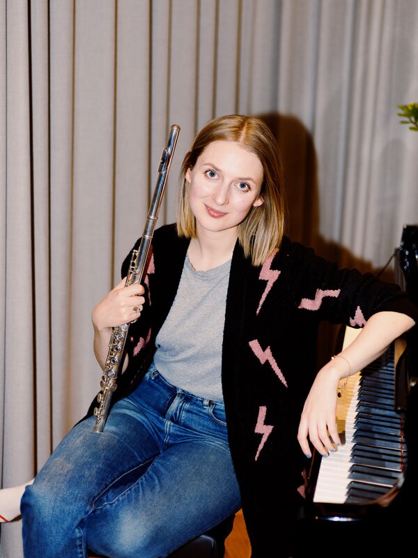 Composer Claire Wickes sits on a piano bench while holding her flute