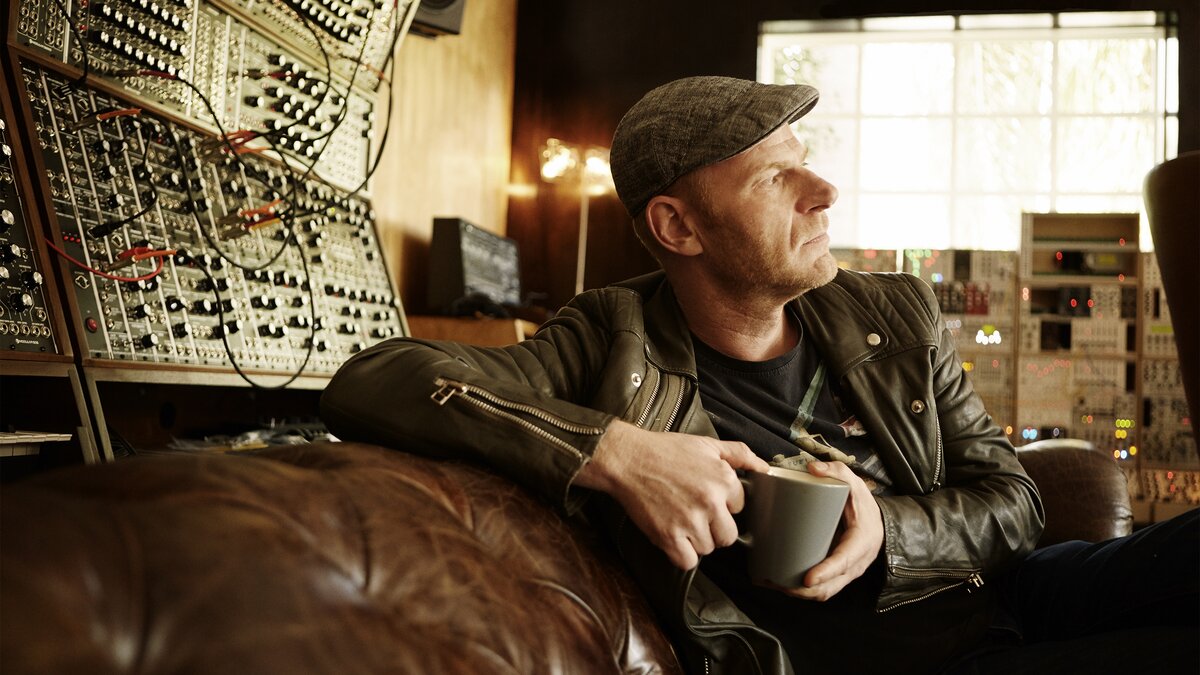 Tom Holkenborg with a mug. There is a large modular synthesizer in the background.